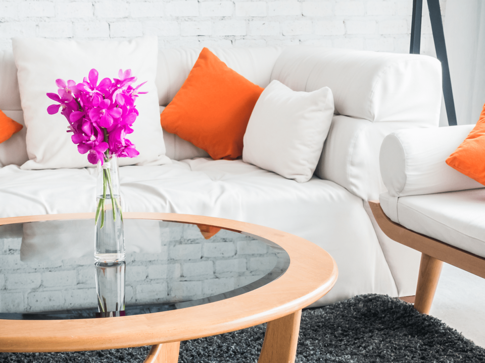 Imagem de uma sala com sofá branco e almofadas brancas e alaranjadas. Uma mesa de madeira com tampo de video ao centro, com um vaso de vidro com flores na cor lilás - flores para a casa